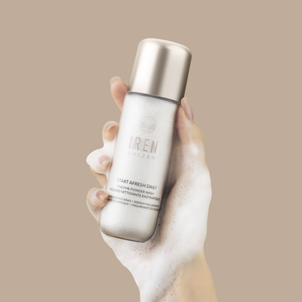 A hand covered in foam holds a bottle of IRÉN Shizen START AFRESH DAILY Enzyme Powder Wash, a gentle rice enzyme cleanser and daily exfoliator, against a plain beige background.