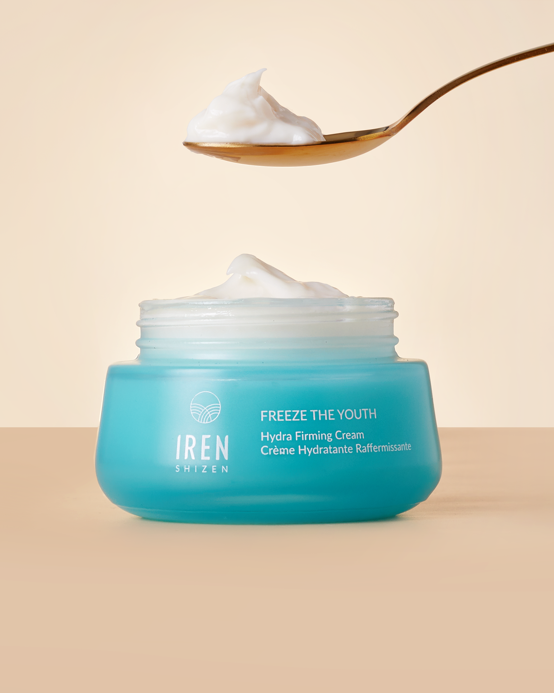 A spoon of cream, infused with grape extract, is suspended above an open jar of FREEZE THE YOUTH Hydra Firming Cream against a beige background. #all-groups