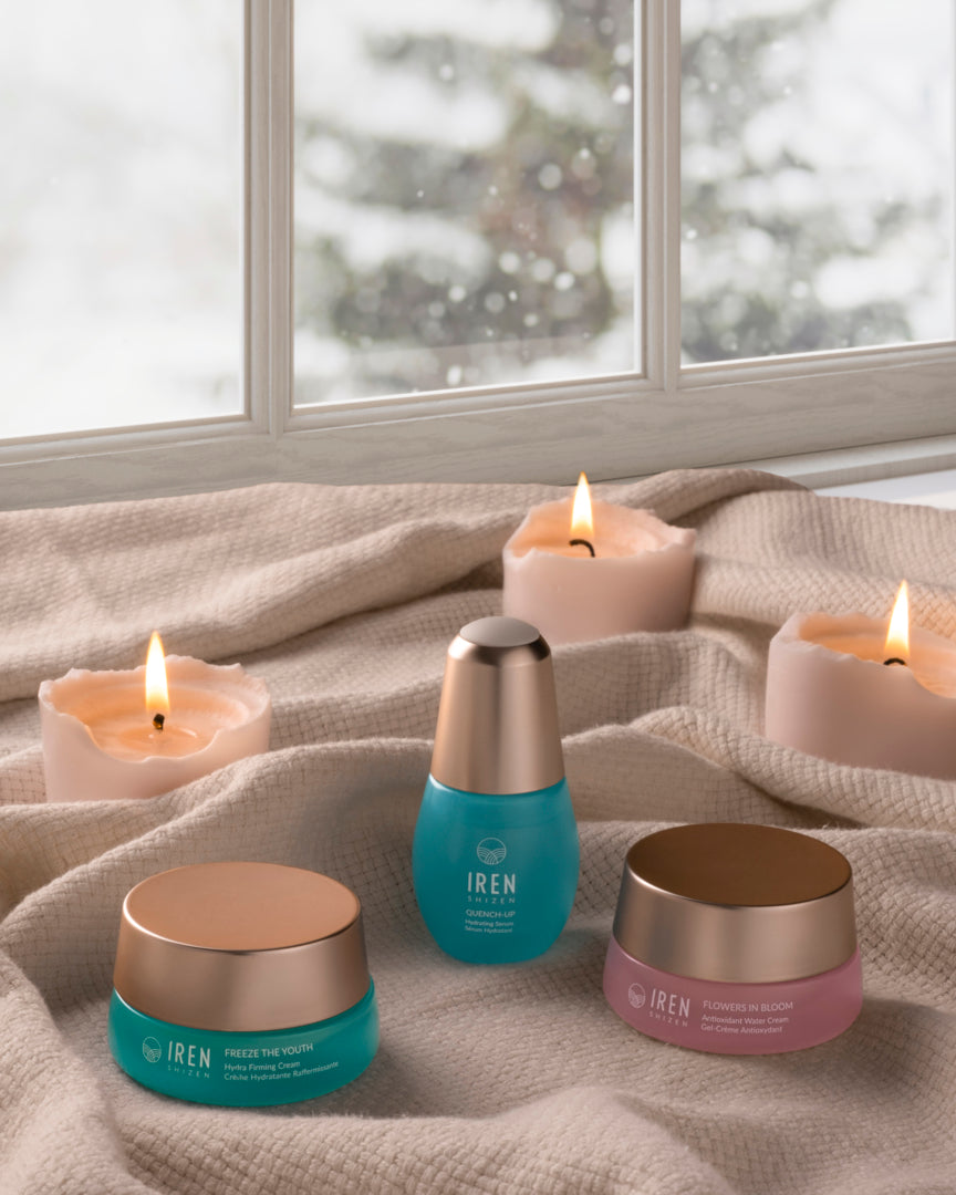 The MOCHI SKIN Instant Glow Travel Set by Fast Bundle and three lit candles are arranged on beige fabric in front of a window with a snowy outdoor view.