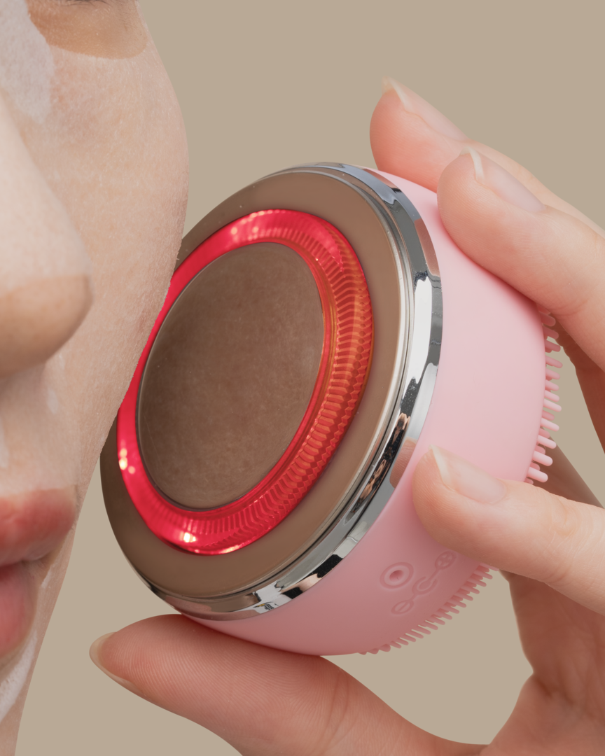 A woman is holding a IREN Shizen SKIN GENIE PRO Cleansing Brush + LED Light Therapy device to her face.