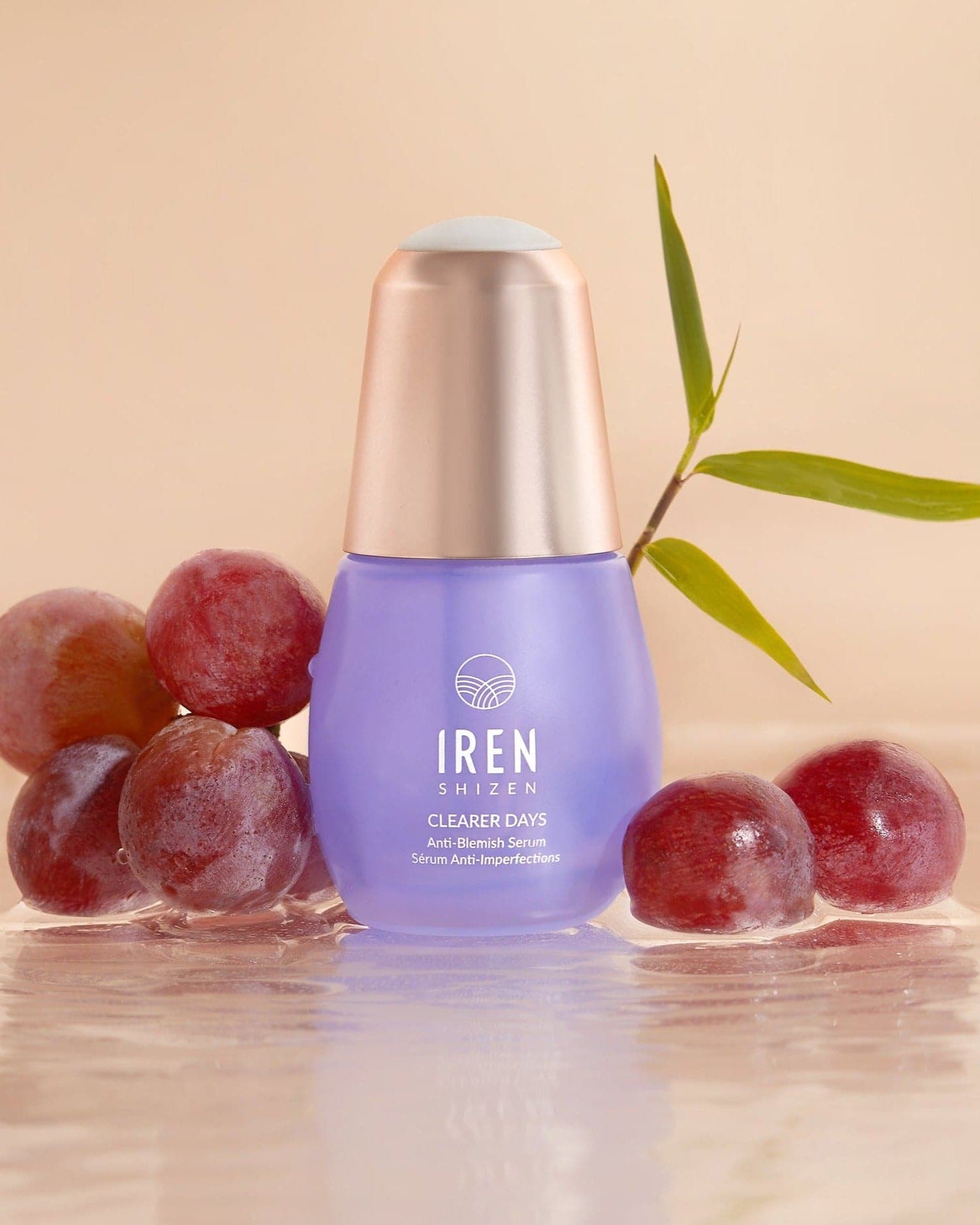 A purple bottle of CLEARER DAYS Anti-Blemish Serum from IREN Shizen is nestled among red grapes and a bamboo leaf on a reflective surface, showcasing its prowess as an anti-blemish solution.
