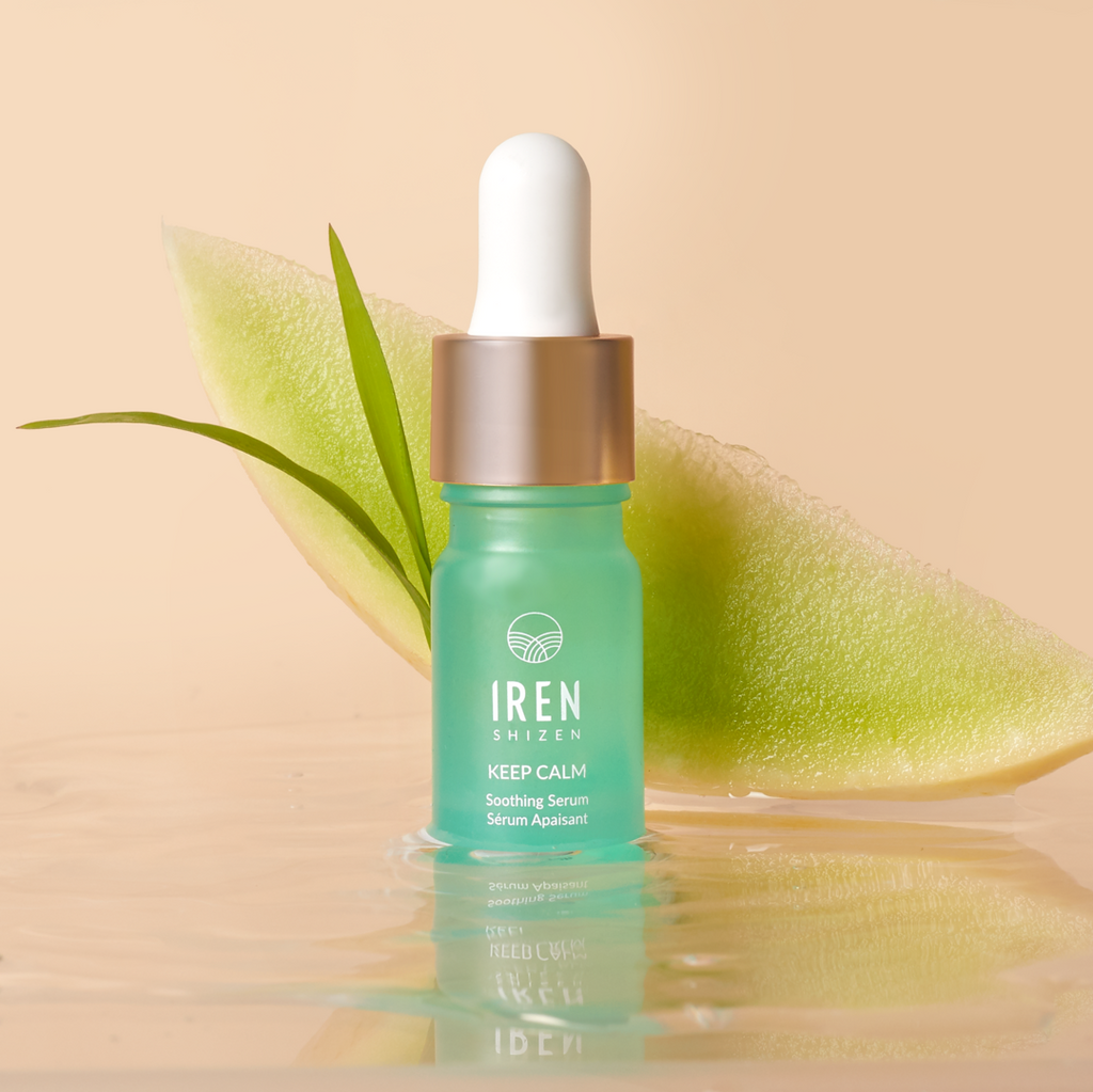 A green glass bottle of IREN Shizen AFTER SUN Repair Routine with a dropper cap is placed in front of a slice of melon and a green leaf, all set against a neutral background—perfect for your after-sun routine.