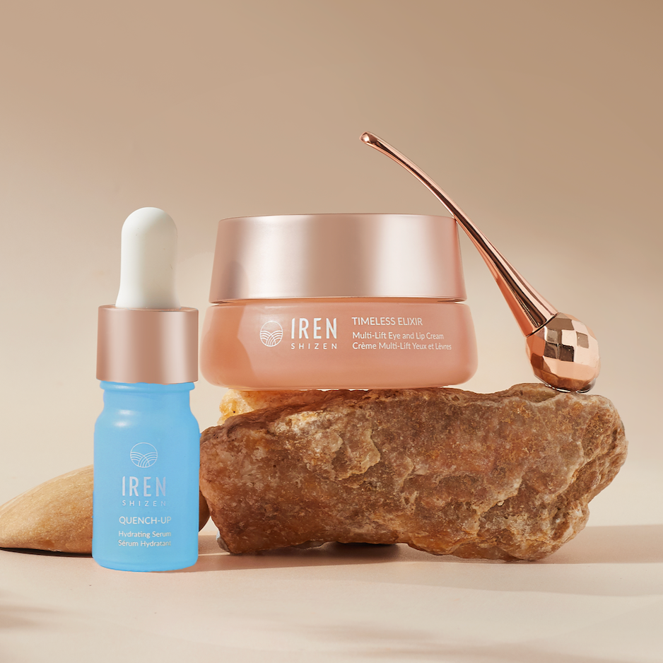 A blue hydrating serum bottle and a pink multi-use lip care cream jar from the IREN Shizen TIMELESS ELIXIR Hydrating Eye Skincare Kit are placed on a rock. A rose gold applicator is also visible, along with an anti-aging eye cream designed to target dark circles. The background features a beige gradient.