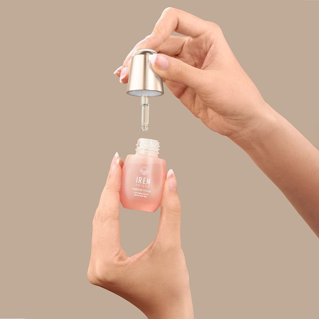 Hands hold a dropper and a small pink bottle from the CARE IN PAIRS Valentine's Day Duo Set by Fast Bundle—an essential part of any skincare routine—against a beige background.