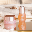 The CARE IN PAIRS Valentine's Day Duo Set by Fast Bundle, featuring a moisturizing cream and revitalizing serum in elegant pink and silver packaging, rests on a reflective surface. Soft fabric and dried flowers in the backdrop create a serene setting for enhancing your skincare routine.