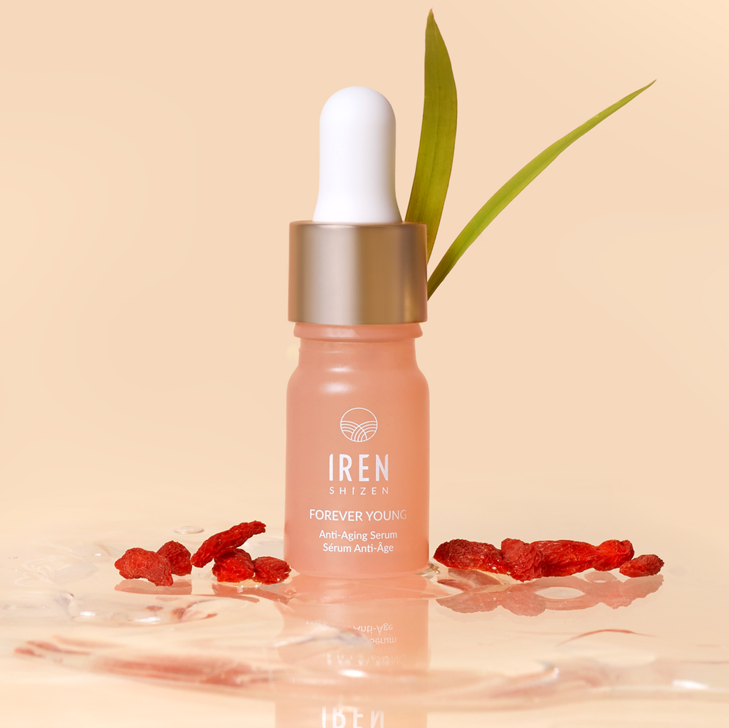 A bottle of IREN Shizen's "TIMELESS ELIXIR Anti-Aging Eye Skincare Kit" is displayed with Goji berries and a green leaf in the background on a light peach surface, perfect for targeting the eye area and reducing dark circles.