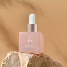 A bottle of the IREN Shizen PINK OCTOBER Skincare Set's Bloom of Youth Restoring Facial Oil, designed for Breast Cancer Awareness and featuring a dropper cap, is placed on a rocky surface against a beige background.