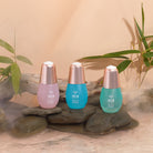 The IREN Shizen 3-SERUM ROUTINE Custom Set, in pastel pink, blue, and turquoise bottles, rests on smooth rocks with bamboo stems and driftwood in the background.