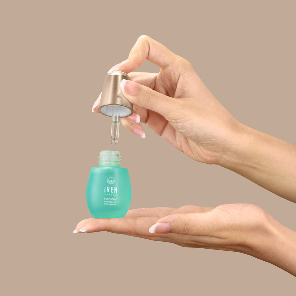 A hand is holding a dropper above a small blue AFTER SUN Repair Routine bottle by IREN Shizen, releasing a droplet into an open hand below.