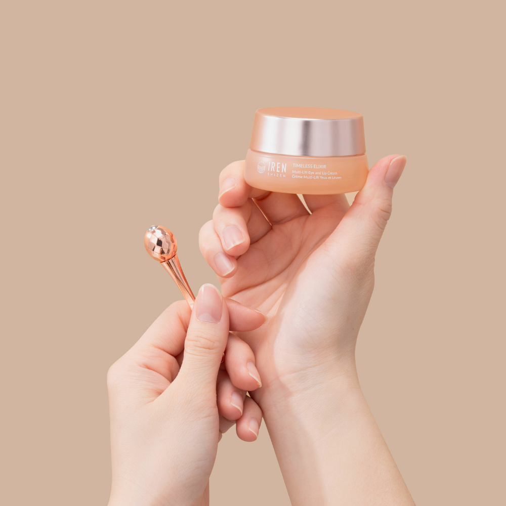 A person holds IREN Shizen's TIMELESS ELIXIR Multi-Lift Eye and Lip Cream in one hand and a rose gold applicator in the other, showcasing the anti-aging formula against a plain beige background.